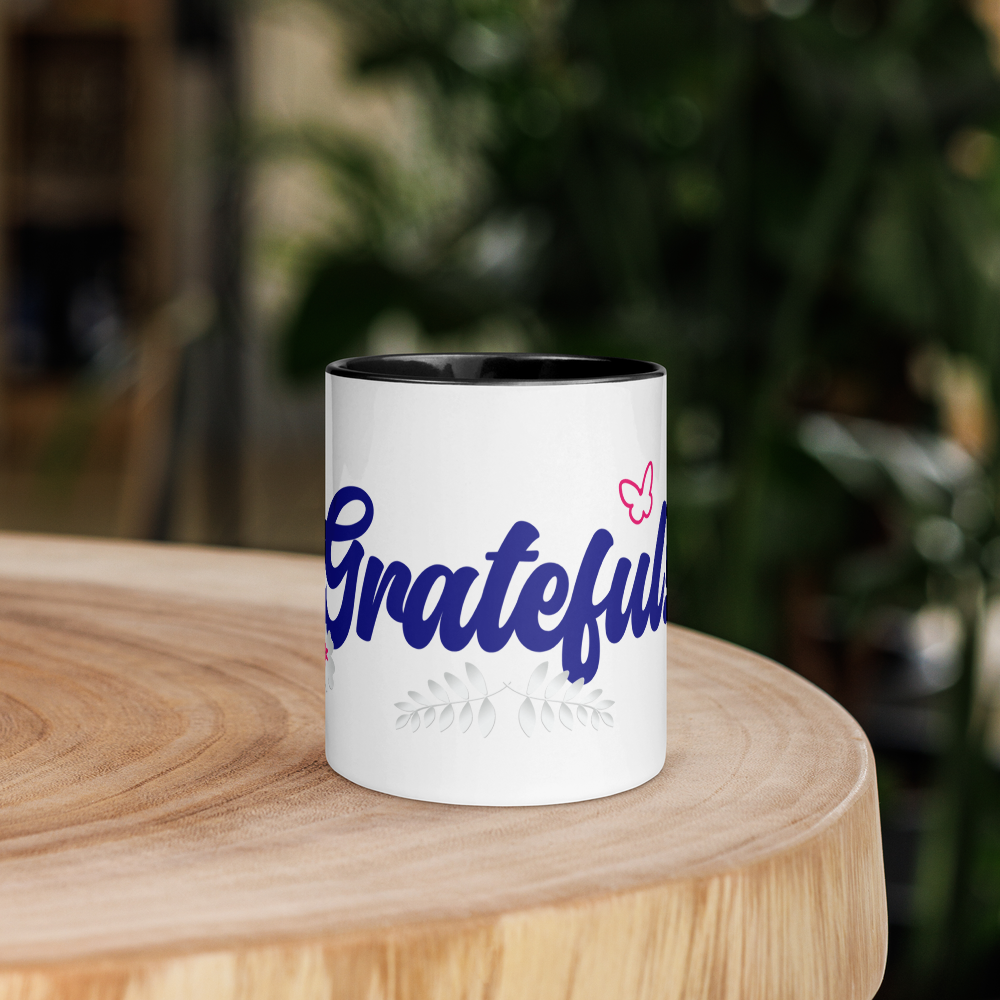 A white ceramic mug with the text 'Grateful' written in a blue, decorative font. The mug has a colorful rim, handle, and interior.